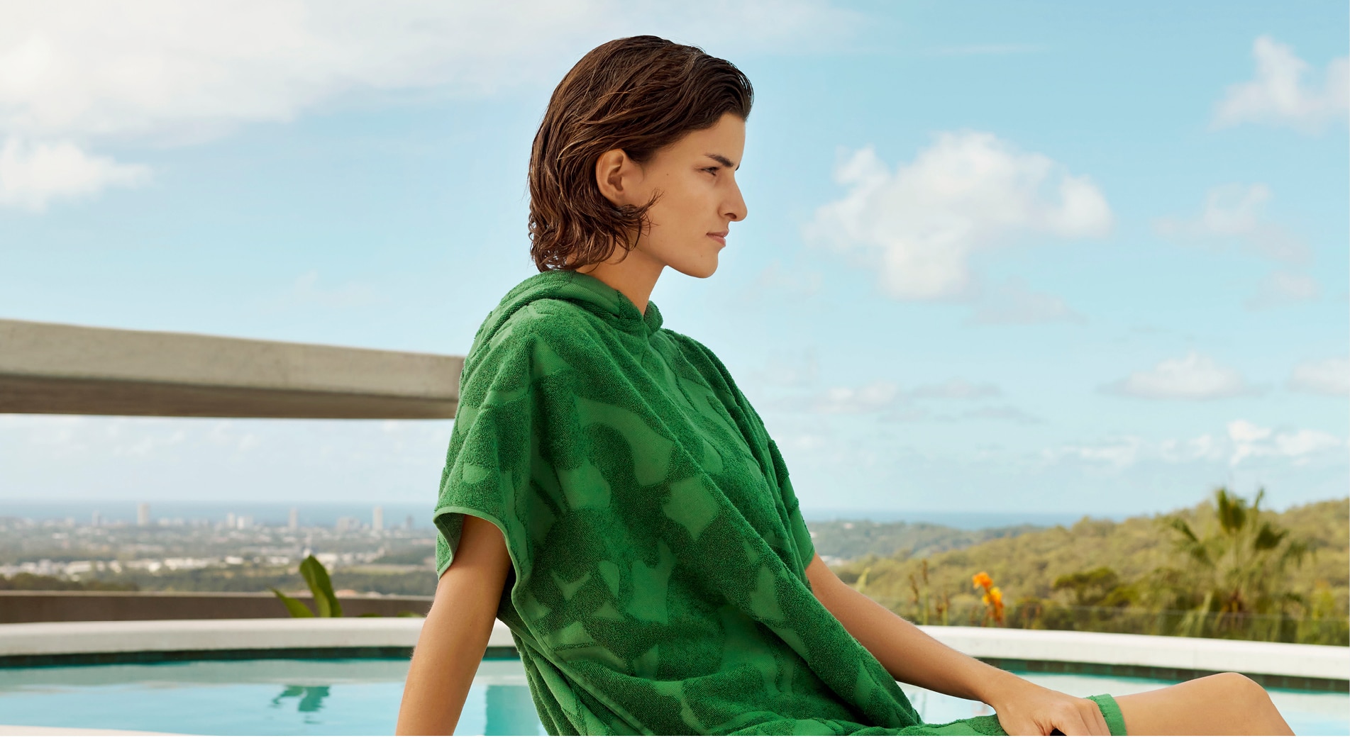 white woman with short brown hair sits in front of a pool, wearing a palm green hooded poncho with botanical design.