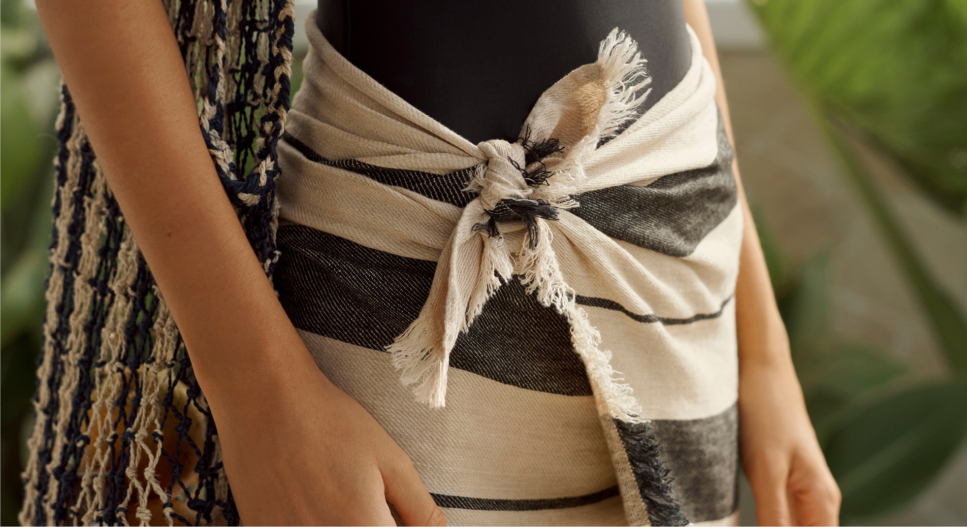 a close up shot of a white woman wearing a sarong as a skirt. it is tied on a knot at her waist. she also has a striped bag, slung over her shoulder resting against her hip. blurry green plants in the background.