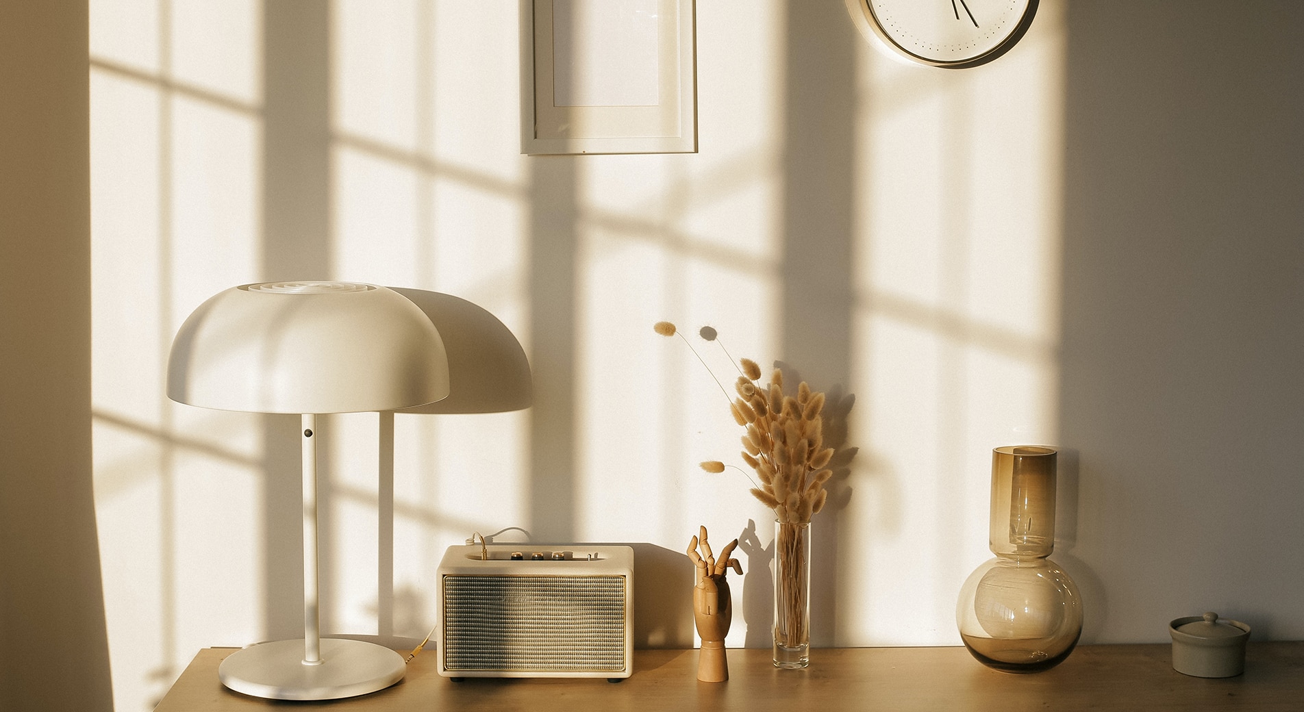 better sleep tips. image taken of someones desk, with natural light coming in through window. on the desk is a white lamp, old radio, dried flowers and a glass vase. a framed image and clock hang on the wall. all in neutral colours.