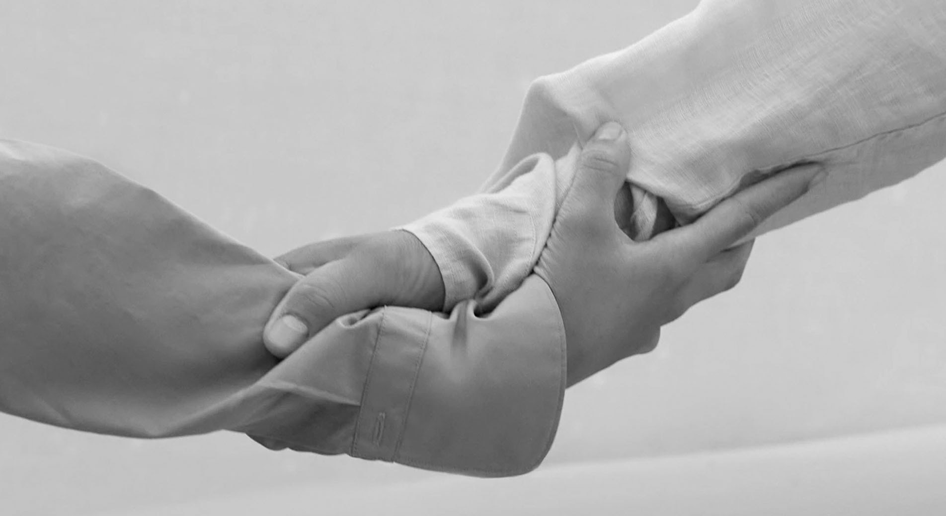 black and white shot of two forearms, in long sleeve button up shirts. their hands are clutching onto the others' wrist, in a supportive gesture.