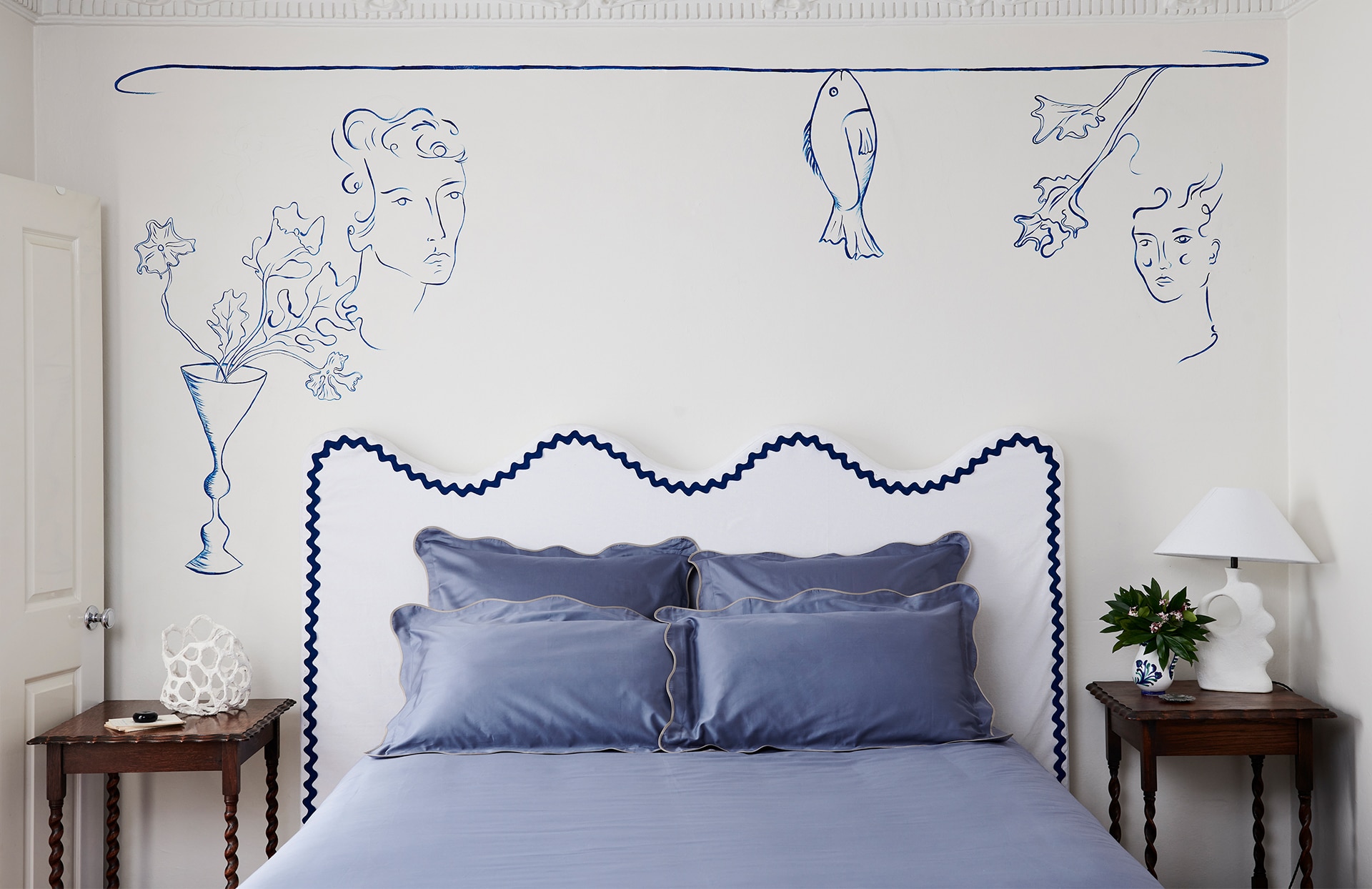 landscape image of bailey jones master bedroom. bedding made with sheridan tamber collection in smokey blue, with scalloped edges. scalloped white bedframe with blue trim behind. wooden side tables on each side of bed. white wall has hand painted mural.