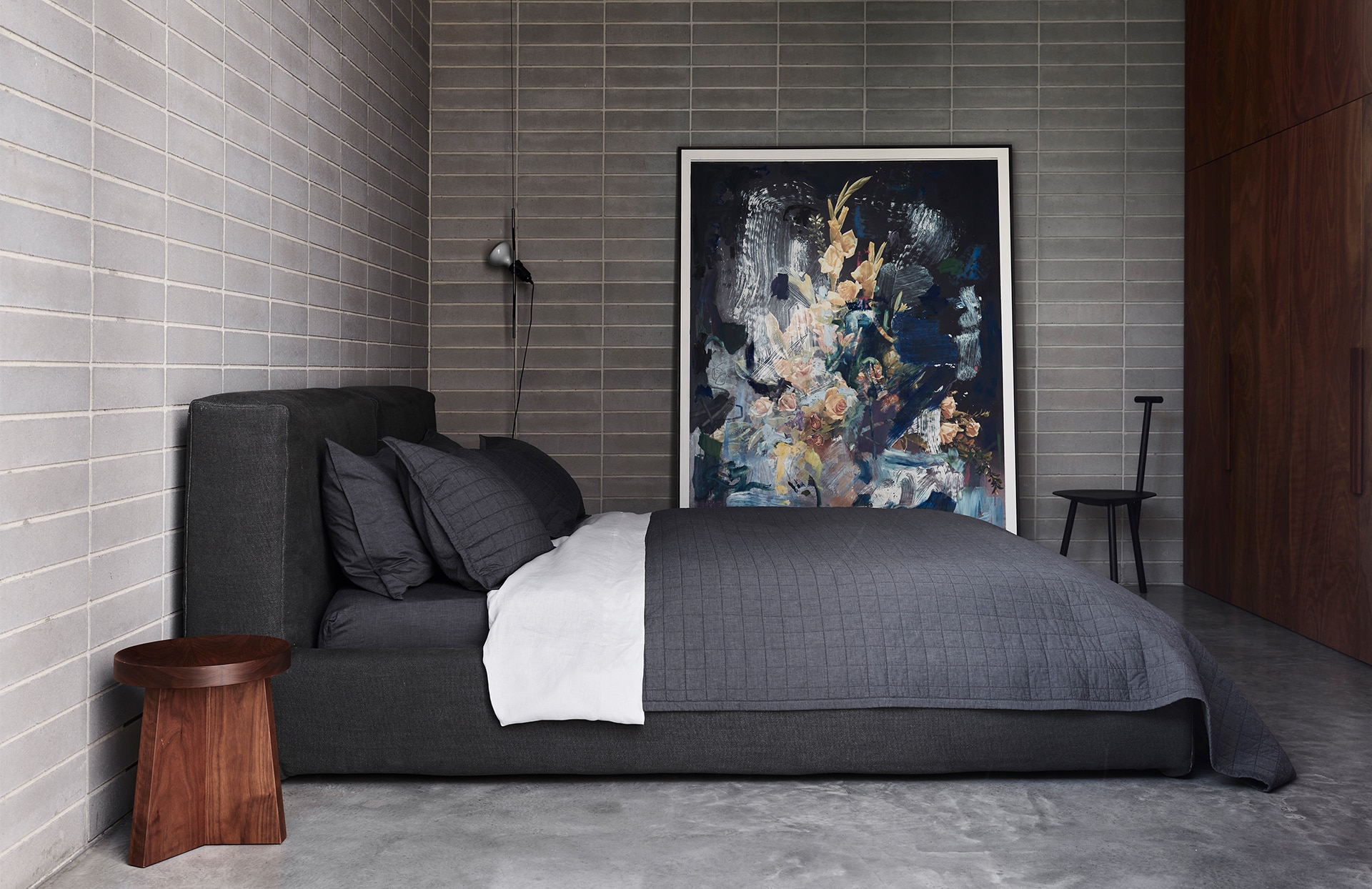 landscape image of james vivian master bedroom. textured bed frame with made up bed. carbon coloured sheeting, pillowcases and bec cover with silver linen sheet. exposed brickwork walls, with large, abstract painting leaning against one wall.