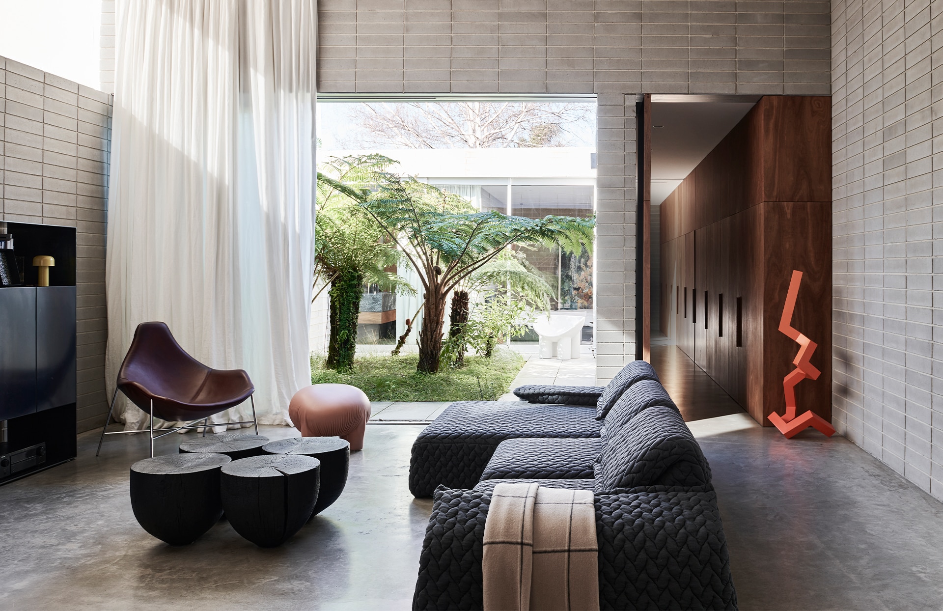 landscape image of james vivian lounge room. long textured couch in foreground, with sheridan check westberry throw over arm. artfully scattered chairs and wooden tables throughout, and abstract orange sculpture. open space leading to garden.