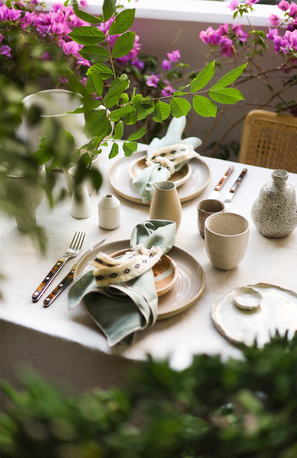 shot through green leaves and flowers, a full table is set up for a dinner party, with abbotson linen napkins, stacked plates, cutlery and glassware. 