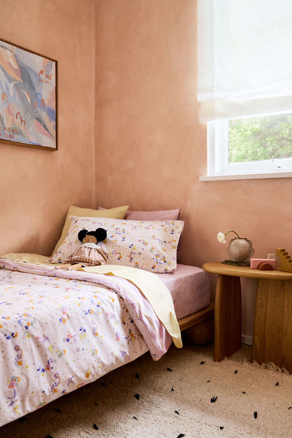 A kids' bedroom. A single bed sits in front of a window, made up with pink and yellow sheets. Next to the bed is a side table with various toys.