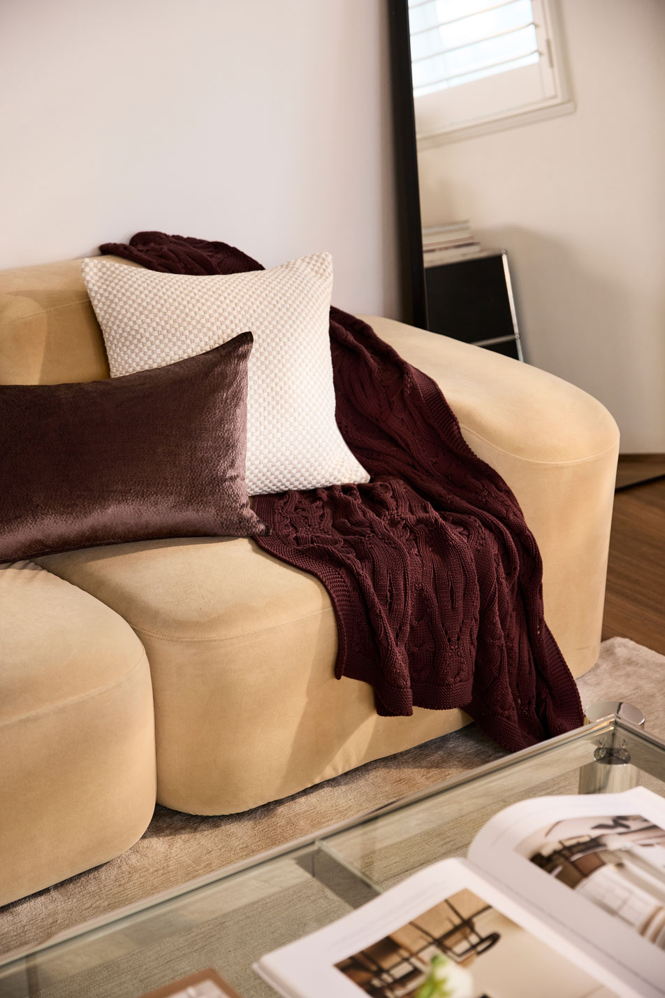 A modern, beige velvet sofa styled with a knit throw and two decorative cushions. In front of the sofa is a glass coffee table with a magazine on top.