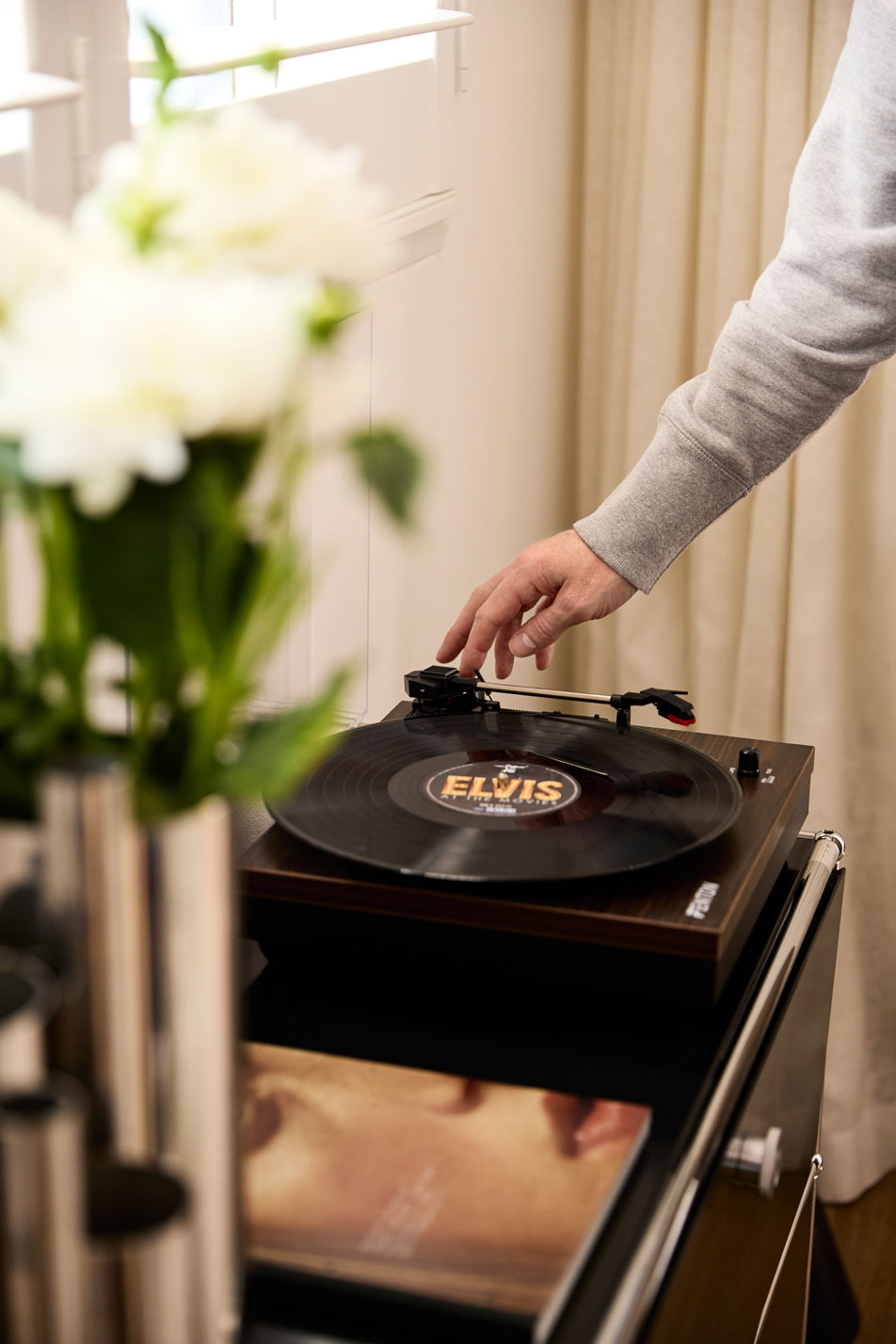 A record player sitting on top of a side table. A man's hand is reaching towards it. Next to the turntable is a magazine and a silver vase with white flowers.