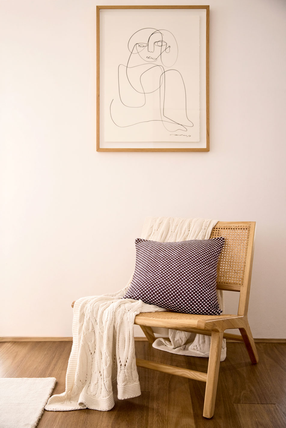 A knit throw and decorative cushion draped over a rattan chair. Behind the chai a framed abstract drawing hangs on the wall.