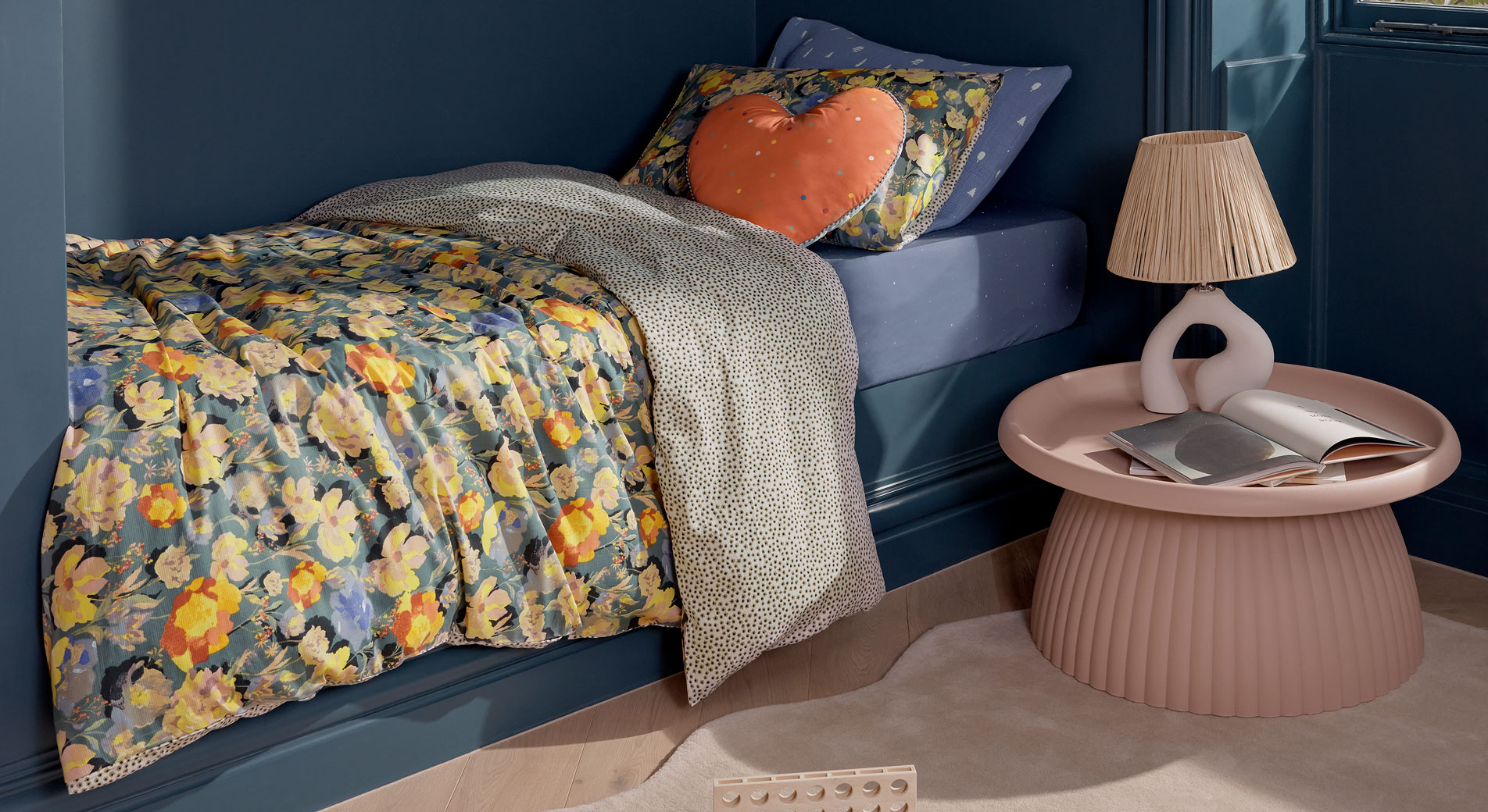 Tips for Transitioning Your Kid to a Big Kids Bed, photo of the corner of a kids bedroom, a kids bed with floral sheets and heart-shaped cushion next to a pink table with a pink lamp and open books.