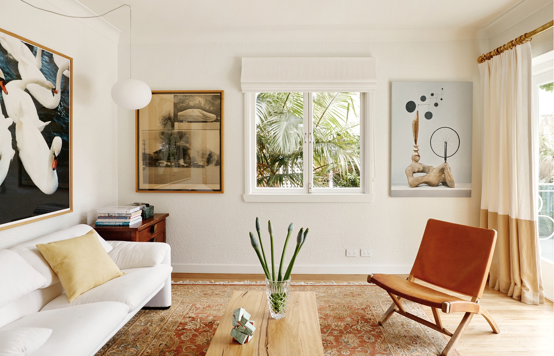 landscape image of pip's living room. left side has white couch with sungold sheridan cushion, above is an artwork of swans. rug on the floor with wooden table and vase on top. right side has leather chair, floorlength curtains and sliding doors.