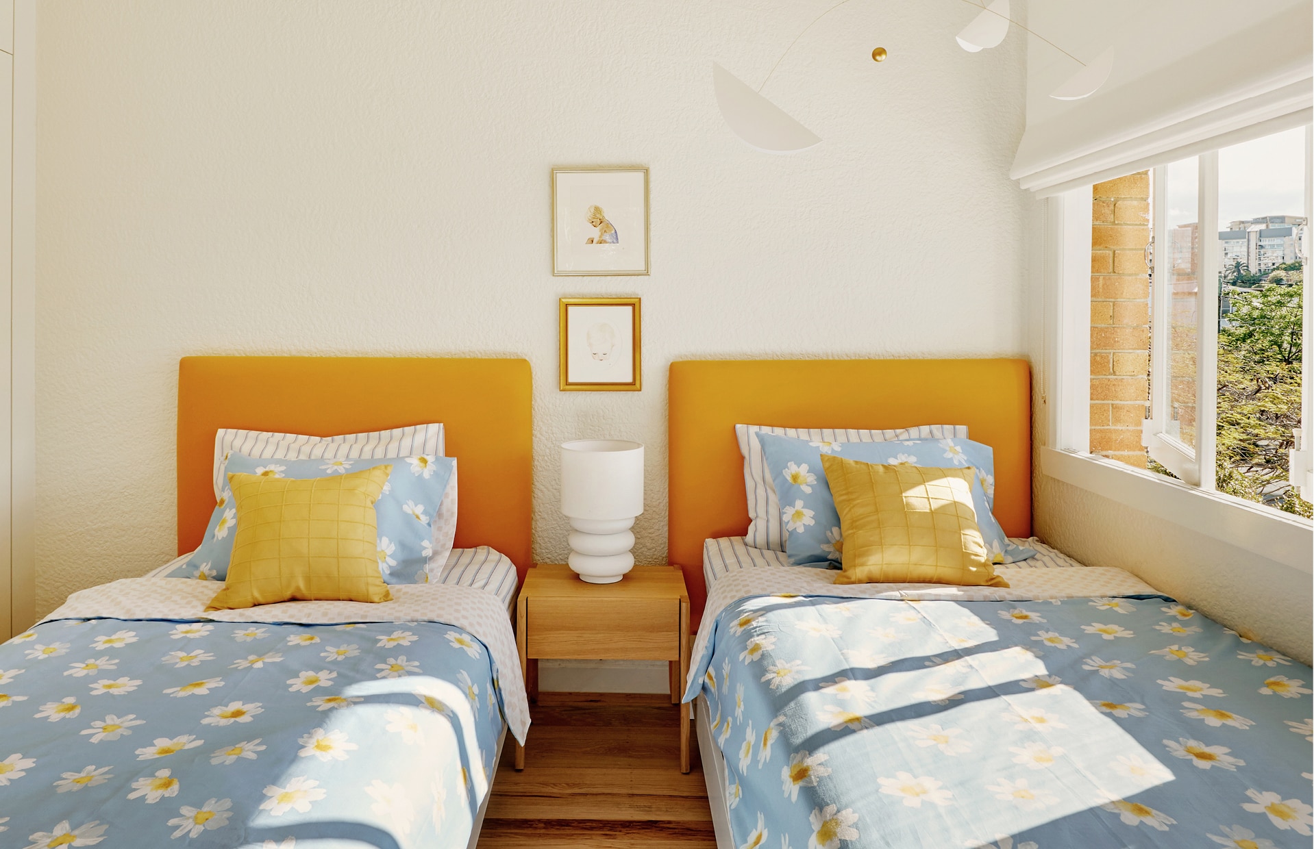 landscape image of pip's kids bedroom. two single beds are styled identially. citrus orange bedhead, striped sheets, and blue quilt cover and pillowcase patterns with daisies. bedside table sits between the beds, with lamp on top. windows to the right.