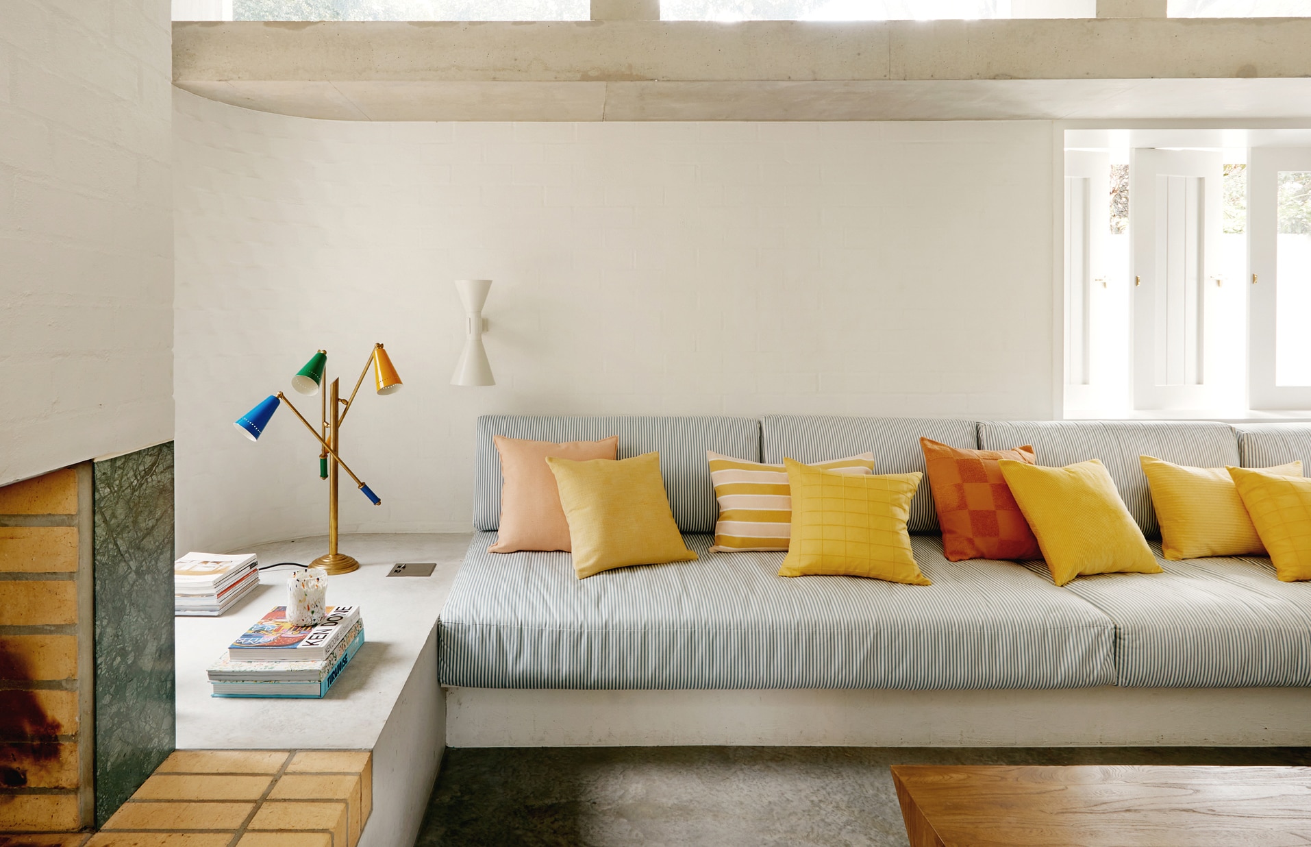 landscape shot of pip's loungeroom. fireplace in foreground, nook behind is stacked with books and multicoloured lamps. lounge is green and white stripes, where multiple sheridan cushions in sunny shades have been styled on top.