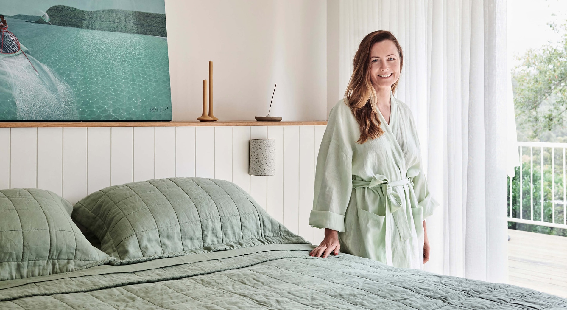 xanthe highfield, white woman with golden brown hair, stand next to her bed. she wears julip linen robe and is smiling, with hand on bed. bed is dressed in cactus abbotson linen.