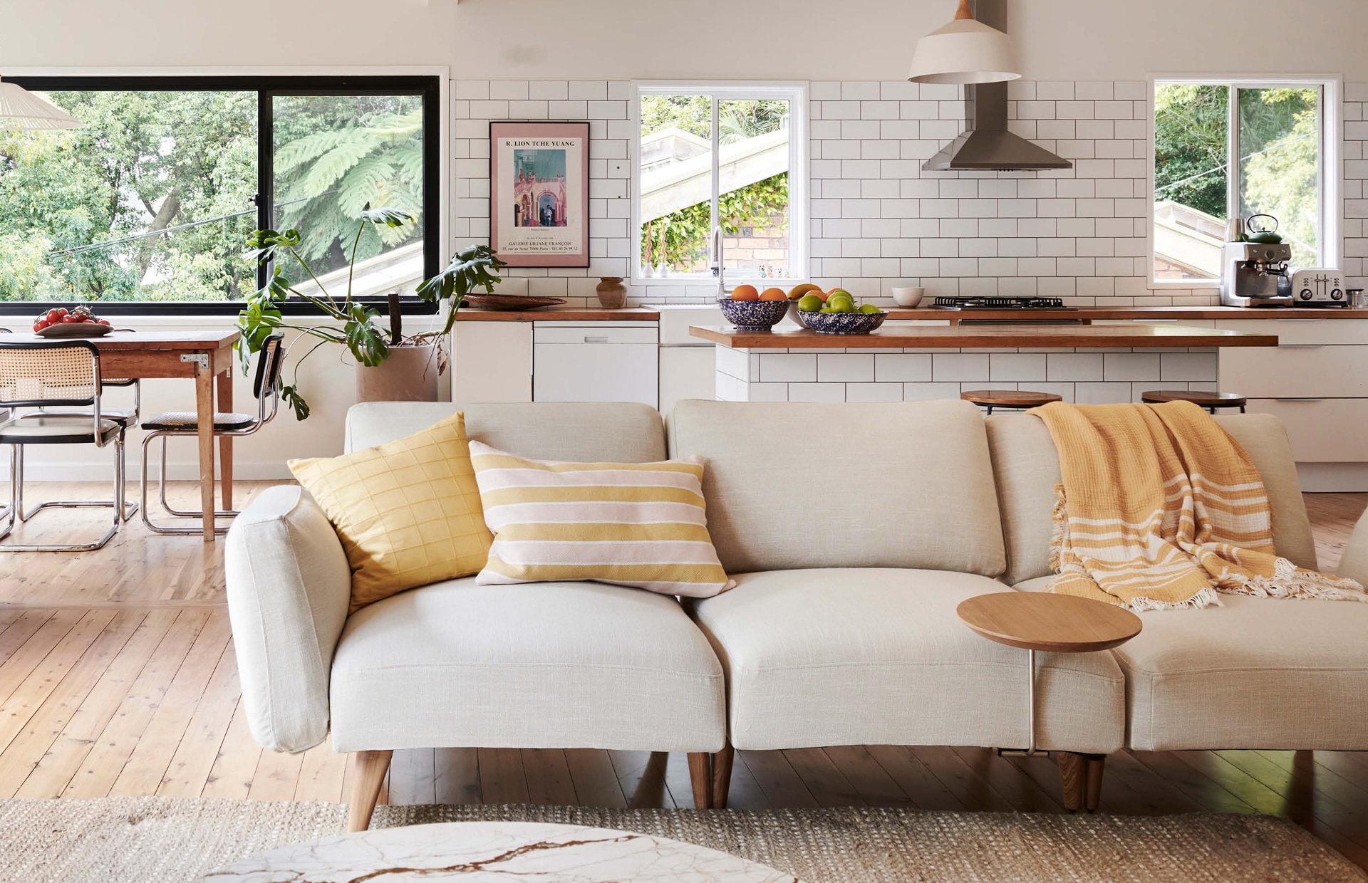 landscape image of xanthe's living room. marble table in foreground, long beige couch stretches out behind it, with yellow cushion, striped yellow and pale pink cushion, and mustard fringed throw decorating it. in background is kitchen and dining table