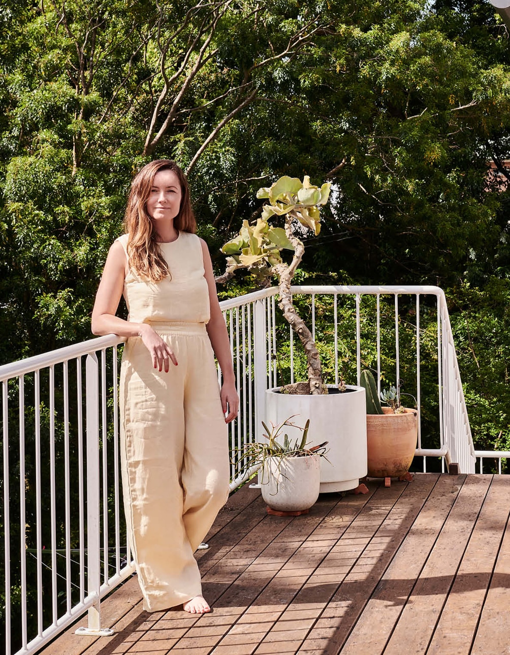 portrait image of xanthe standing on her balcony, resting arm on railing. she wears pastel yellow loungewear and smiles softly at camera. gumtrees and bushland are seen behind her.