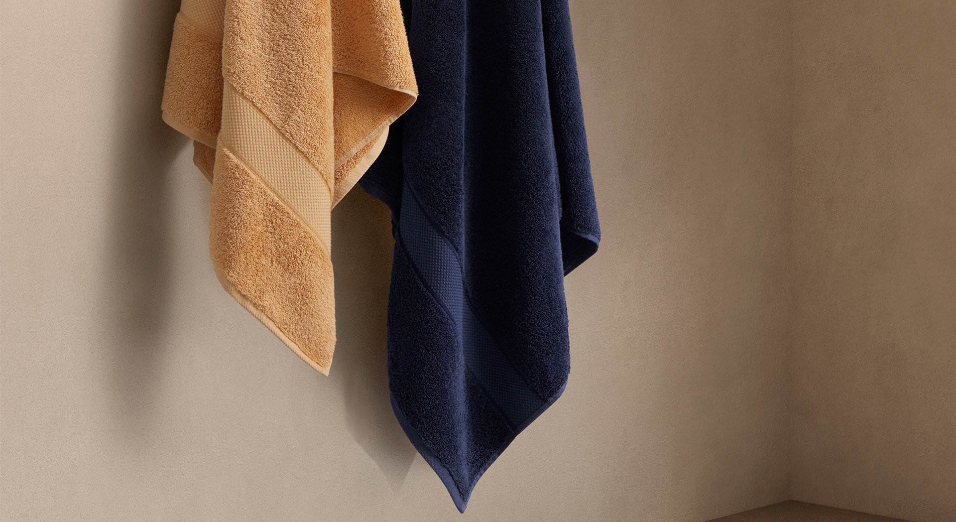 against biege wall, bottom of orange cotton bath towel and navy bath towel hang from hooks.