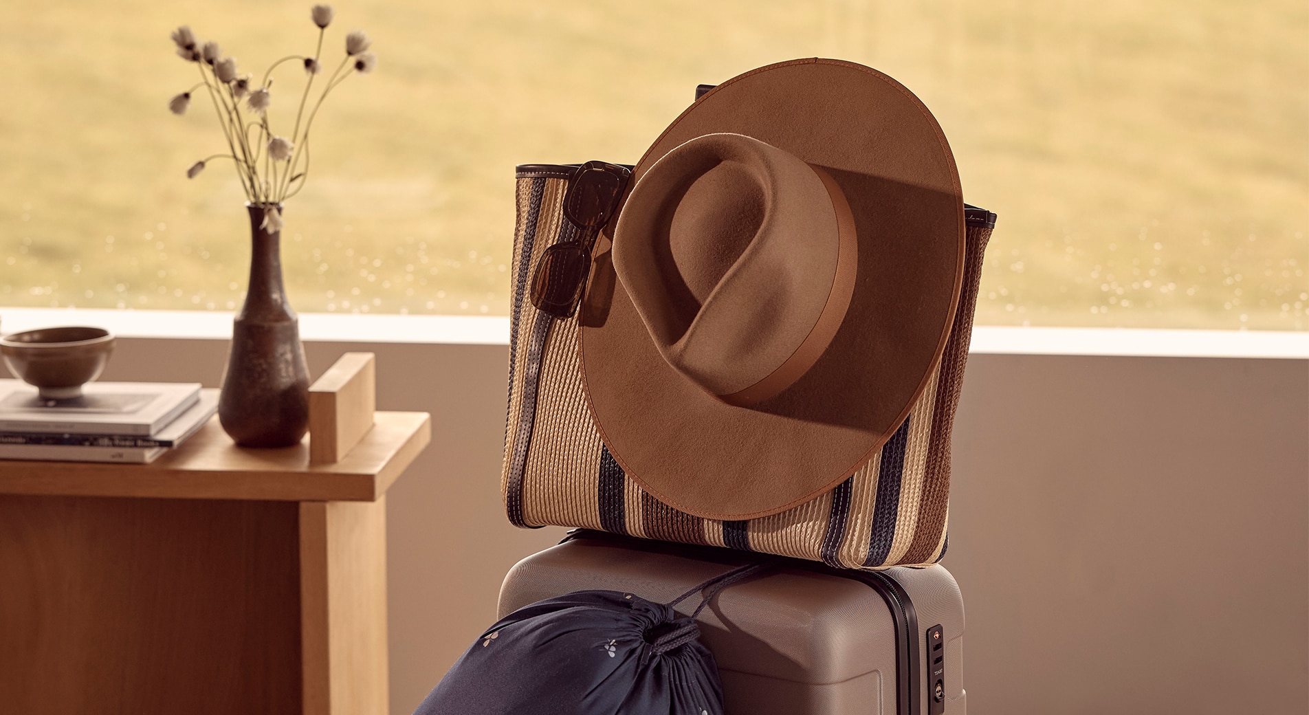 how to make flying more comfortable. suitcase stands on floor, with carry on sat on top and travel blanket hanging off. in the background is wooden desk, out the window is green grass.