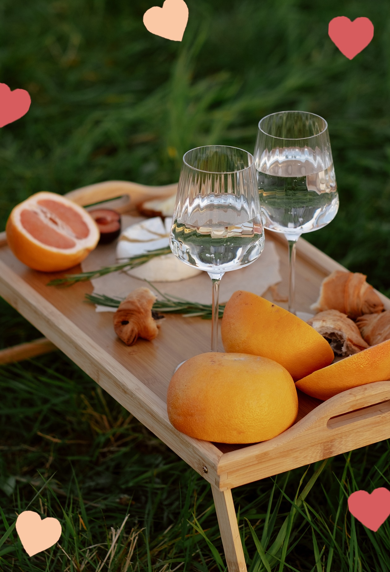 wooden tray table sitting in green grass, with wine glasses, cheese and fruit atop it. scattered hearts decorate the picture.