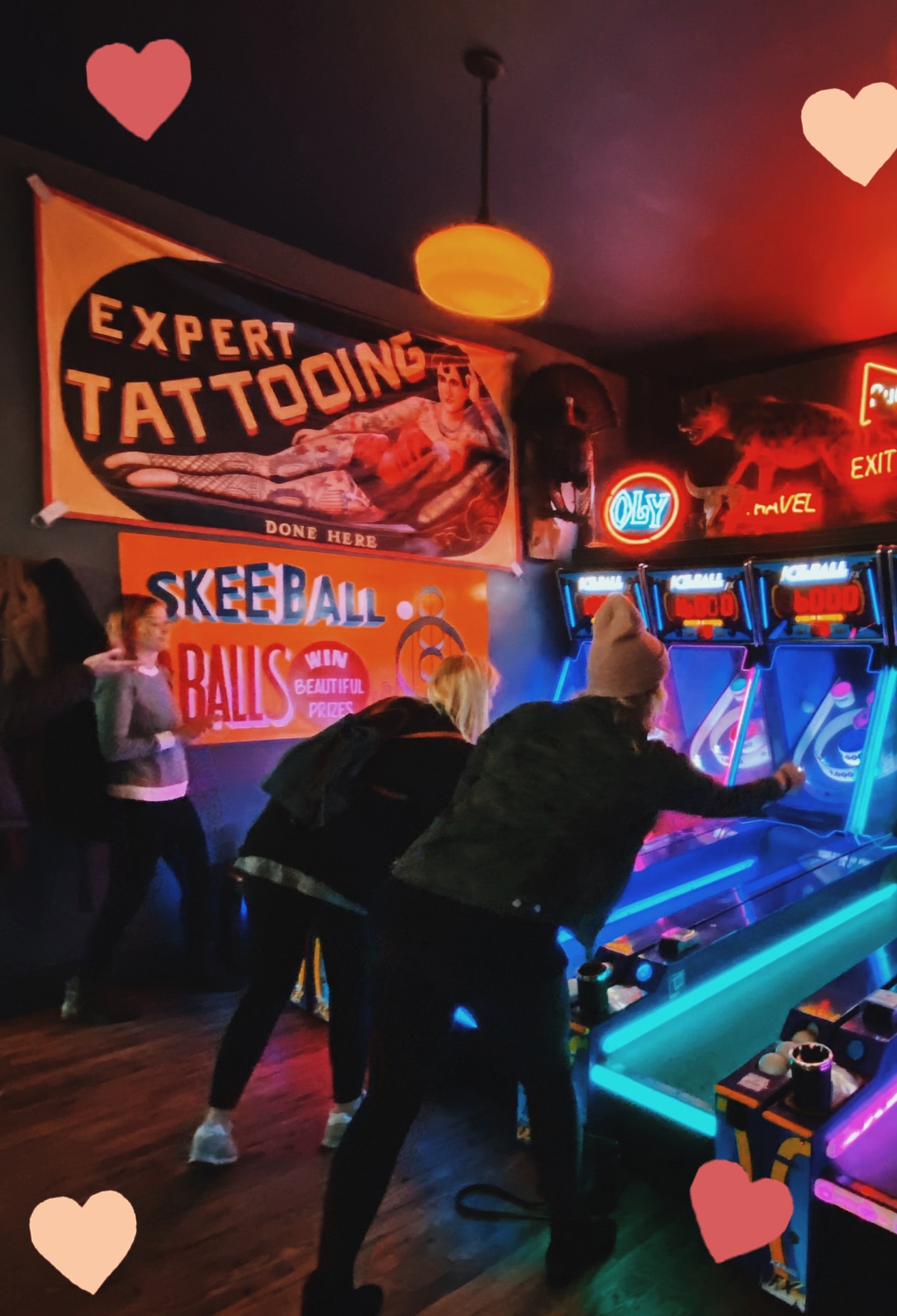 couple plays arcade games in dark arcade, lit by UV lighting from the machines. scattered hearts decorate the picture.