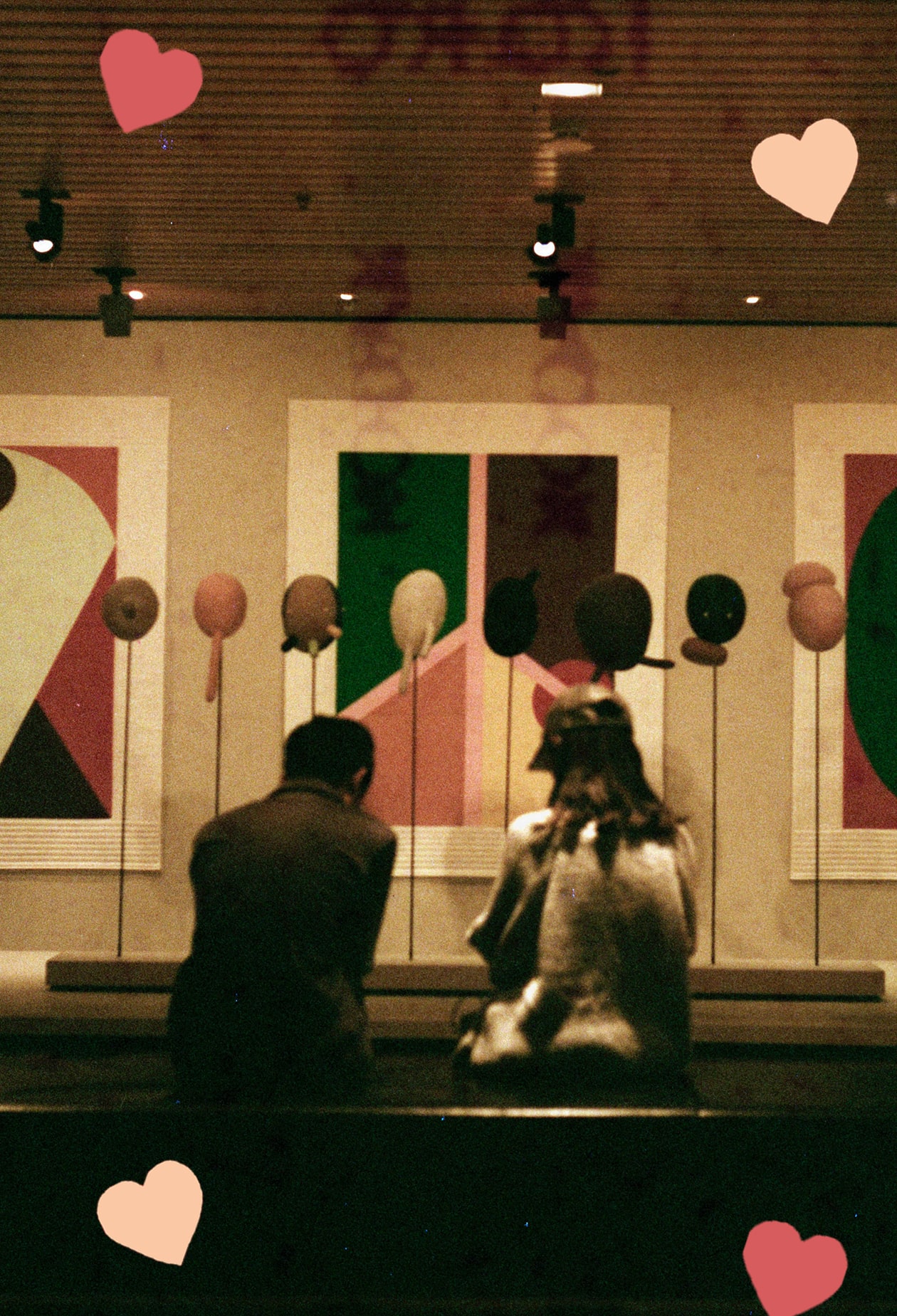 couple stand in front of sculptures and artwork at a gallery. shot on film. scattered hearts decorate the image.