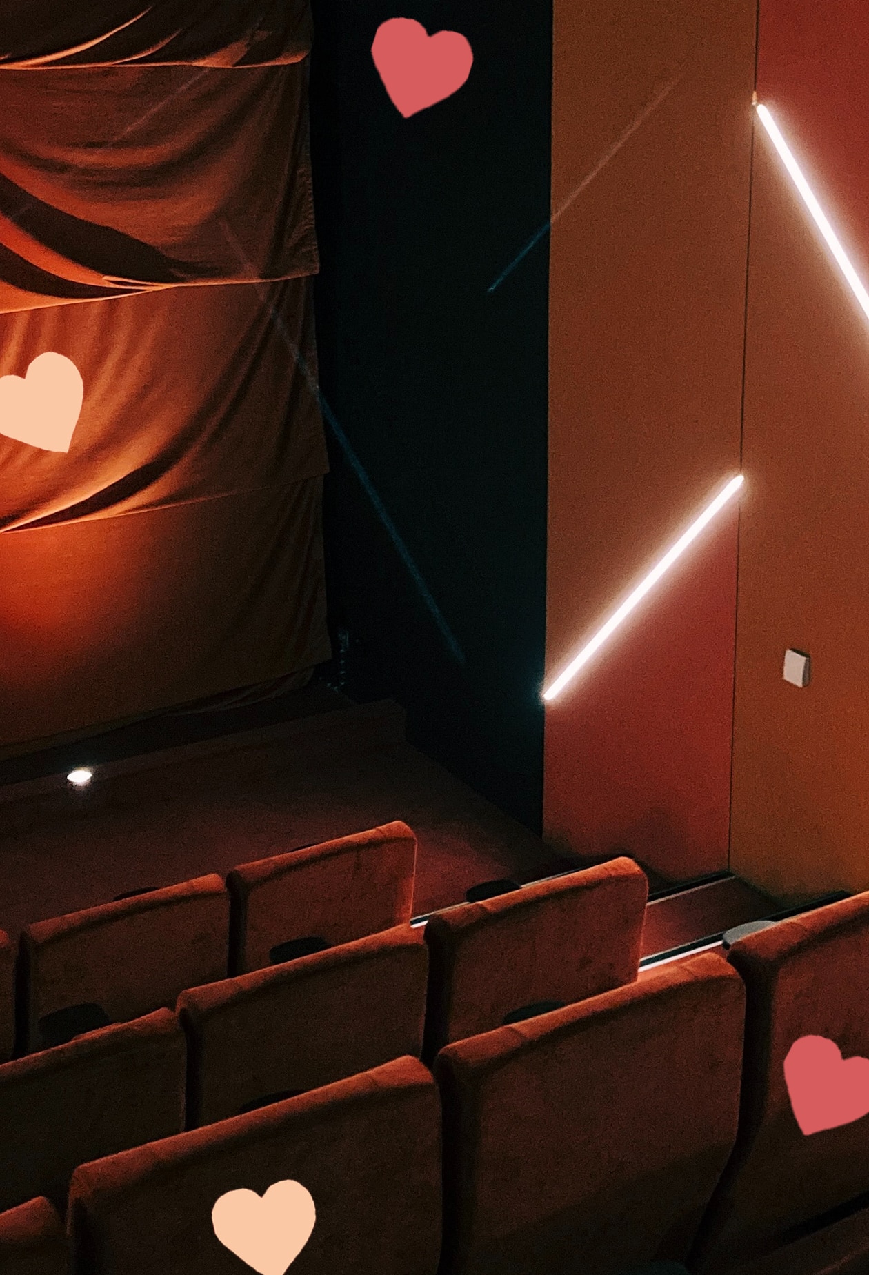 vertical shot of movie theatre, with classic red seats in front, light and curtain in back. scattered hearts decorate the image.