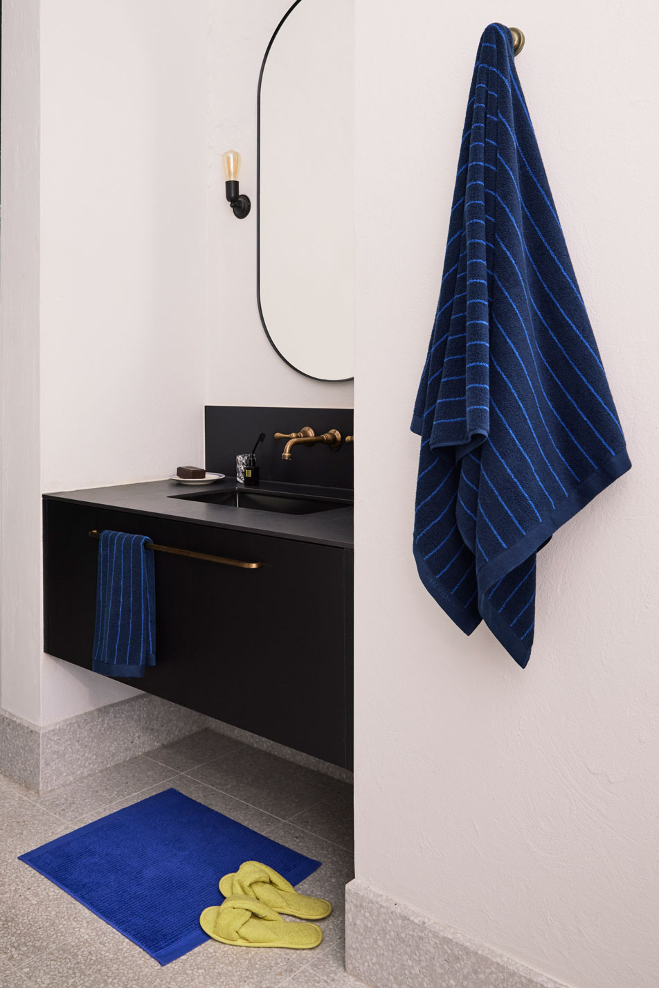 A modern bathroom with a black vanity and oval mirror. On the floor is a small blue bath mat and a pair of yellow slippers. A blue striped towel is hanging from a brass knob on the wall.
