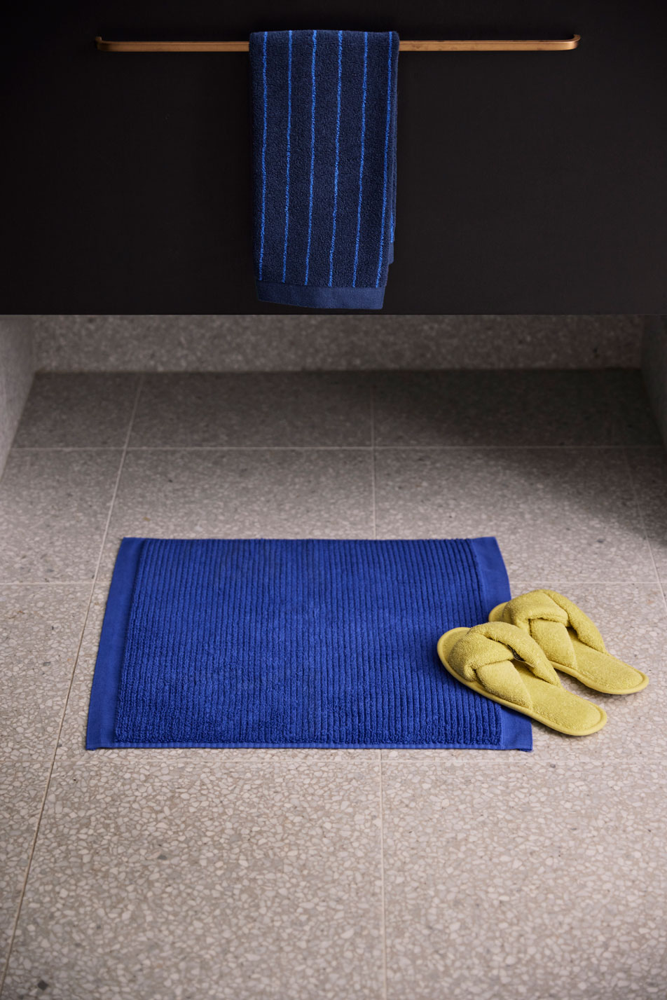 A small blue bath mat with a pair of yellow slippers sits underneath a black vanity with gold hardware and a matching blue hand towel draped over the rail.