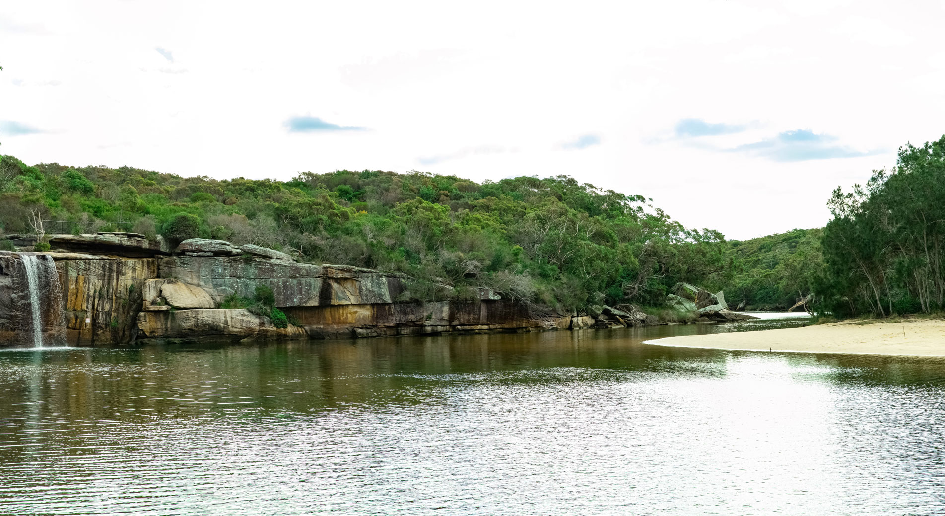 wattamolla lagoon. foreground is light reflected of water, with small waterfall to left side image over rocks. midground has green bushland, background is bright white sky. to the right is a small crop of sand jutting out from bush.