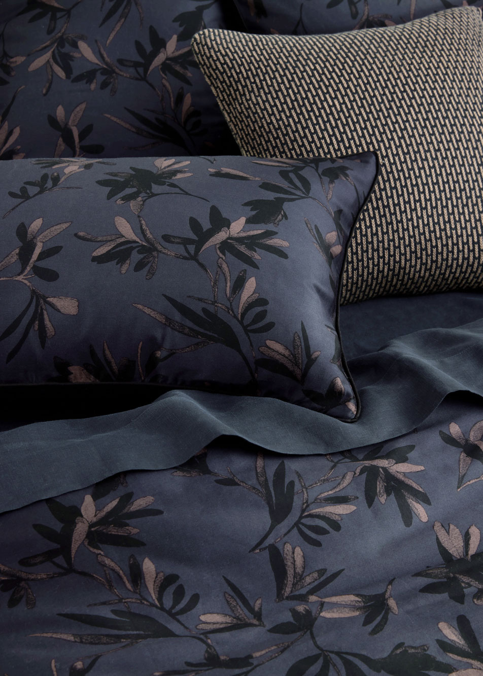 A close-up of a bed dressed in dark bed linen, with black sheets, a black and brown botanical print quilt cover, and a knit cushion.