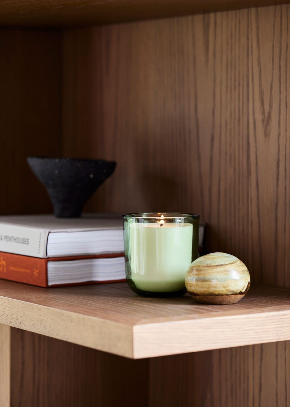 A lit candle sitting on a bookshelf. Next to it is a stack of books and a small ceramic decoration.