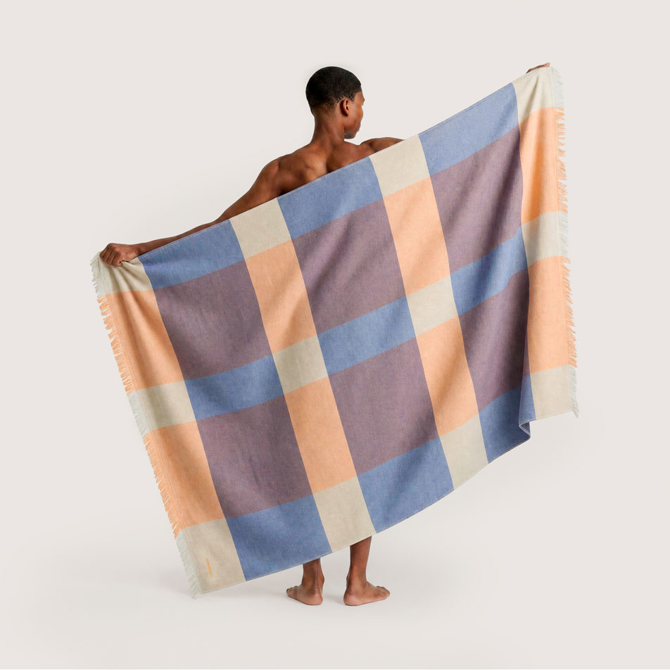 A male model standing with his back to camera, holding up a large checked beach throw in orange, blue and cream