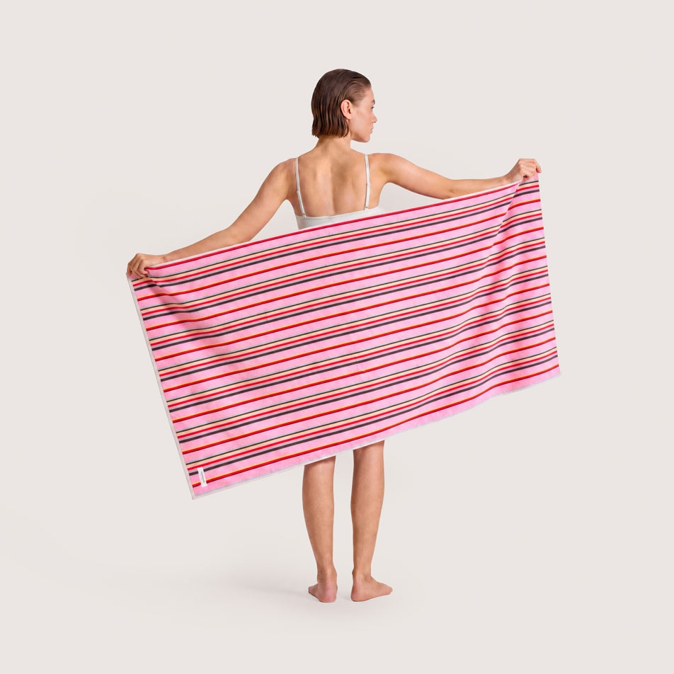 A female model standing with her back to camera, holding up a pink and multicoloured stripe beach towel behind her back