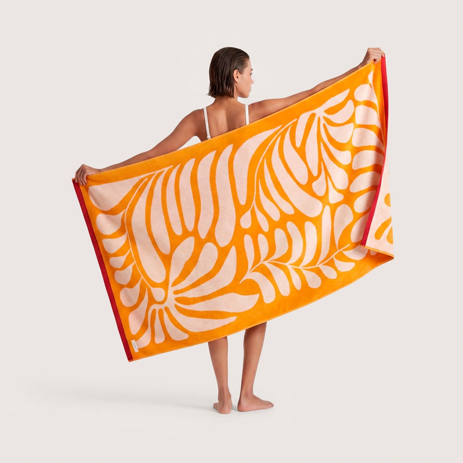 A female model standing with her back to camera, holding up an orange beach towel with a white abstract design on it