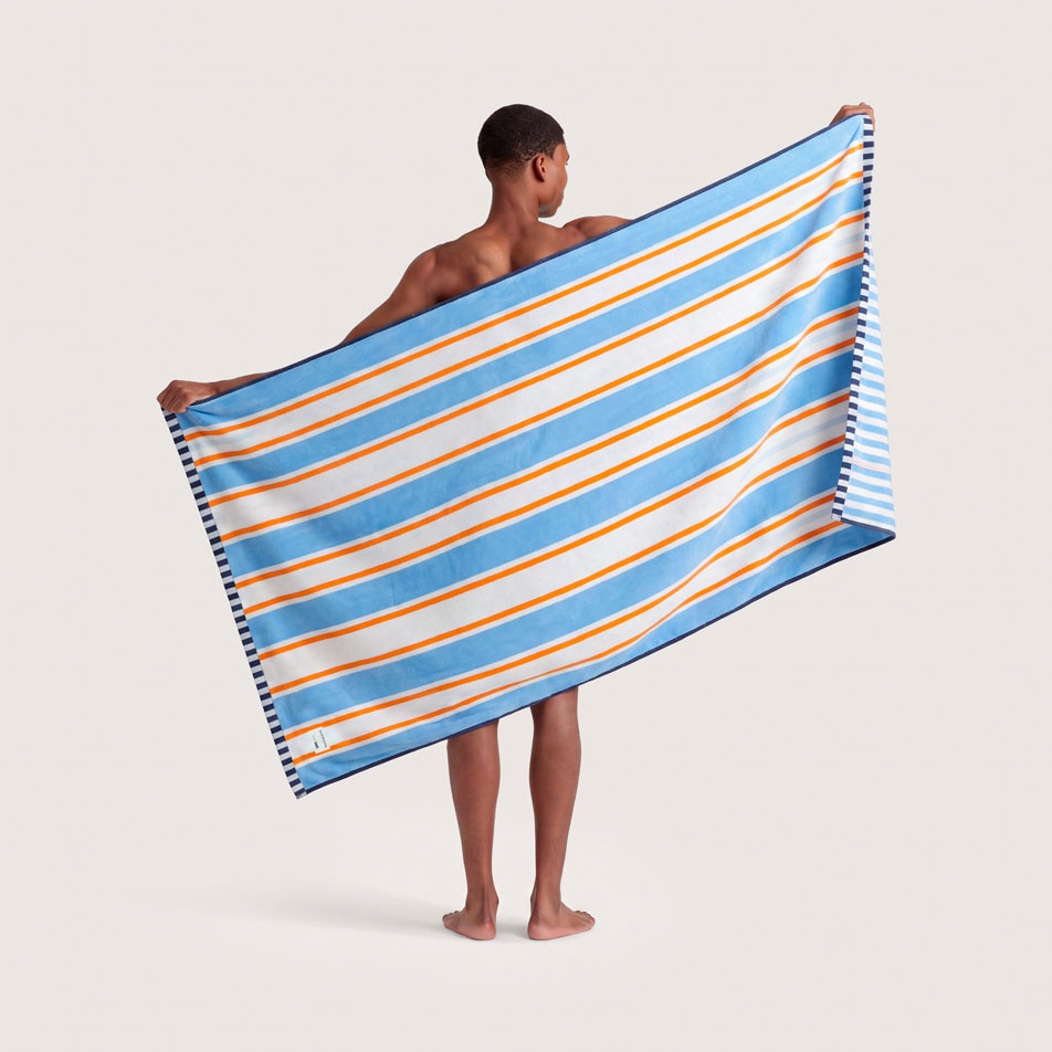 A male model standing with his back to camera, holding up a blue, white and orange striped beach towel behind his back