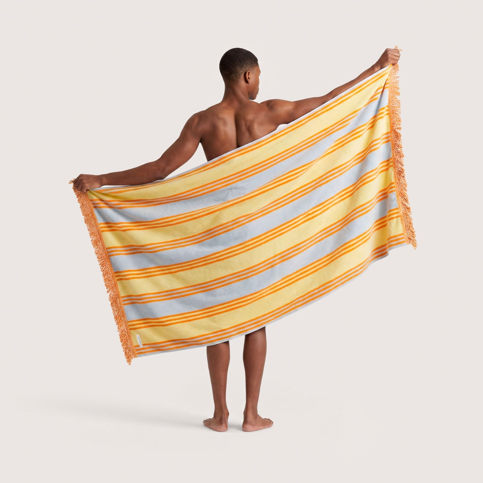 A male model standing with his back to the camera, holding up an orange, yellow and blue striped beach towel