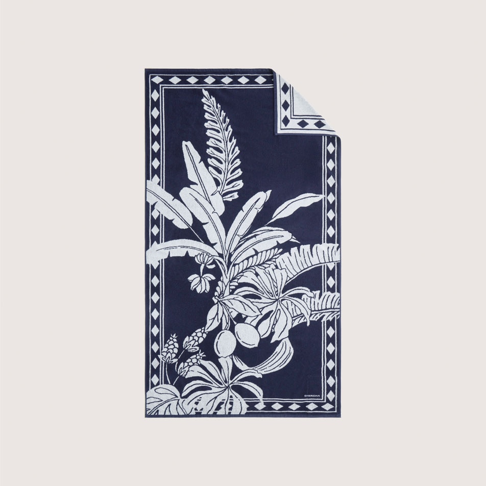 A flat lay image of a navy blue beach towel with a white tropical design on it, featuring banana leaves and tropical fruit